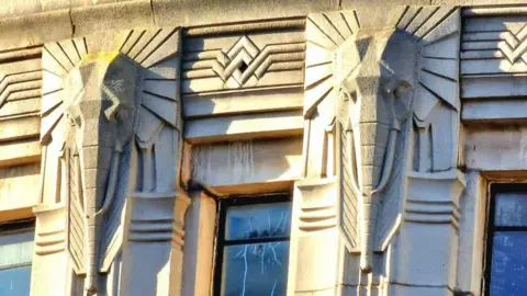 Ian Hughes Ornate stonework in the form of angular, stylised elephant heads surrounding windows with broken glass in them