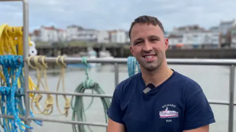A man in a blue t-shirt is standing in front of railings, with a harbour in the background. He has short brown hair and stubble. His T-shirt features a logo of a fishing boat and the name Dylharis. Ropes in blue, yellow and green are attached to the railings.