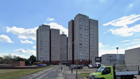 Google Three grey tower blocks in the middle background of the photo. They are about 15 storeys high. There is a patchy road leading up to them and two people are cycling towards the camera. A few cars are parked in front and there are smaller concrete buildings to the right.