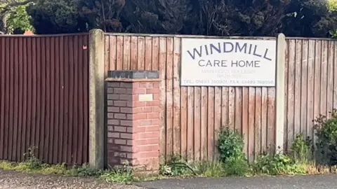 Qays Najm/BBC A brown fence, with a white sign, printed with the words WINDMILL CARE HOME on it.