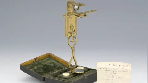 The brass microscope, fully extended. Its base is a small brown box lined with green velvet. There is also a handwritten note beside it in cursive handwriting, the text of which is not legible. 