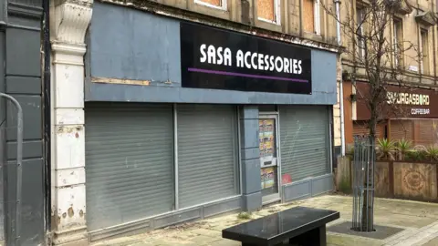A shop front on an urban street. The shop name 'Sasa Accessories' is above the windows. Shutters are down within the shop. A small marble bench sits outside.
