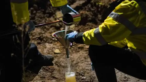 Two workmen in hi-vis jackets sift water into a bottle through a funnel. They appear to be working on a sandy surface.