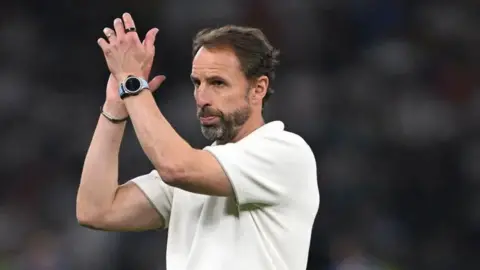 Getty Images Gareth Southgate claps after a football game. he is wearing a white t-shirt and wears a watch and bracelet. he has brown hair and a greying beard