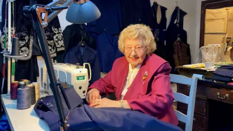 A woman with blond hair and glasses wearing a pink tunic and white blouse sitting behind a sewing machine with some blue material in her hand. 