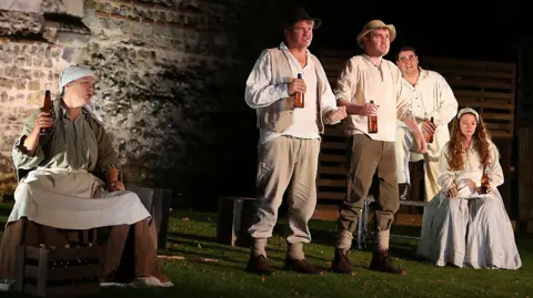 A cast of actors during a performance in front of a brick wall belonging to a castle. On the left, one of the actors is sitting. On the right, three male actors are standing while a female actor sits down. 