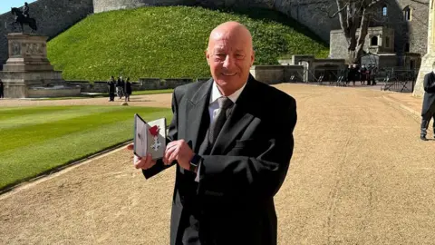 Handout A man in a black suite stands in the grounds of a castle on a sunny day, holding an MBE cross award. He is bald and smiling. Behind him are ornamental gardens and he is standing on a stony path.