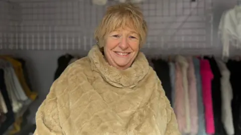 Mary Taylor - a woman with blond, short hair wearing a bright fur poncho smiles at the camera. In the background are a number of colorful garments on clothes rails.