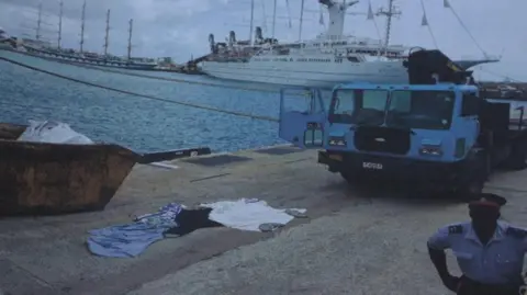 supplied by Chris Purkess A pile of clothes lies on a quayside near a skip and a heavy truck. A cruise ship is in the background and a uniformed officer stands near the camera-taker.