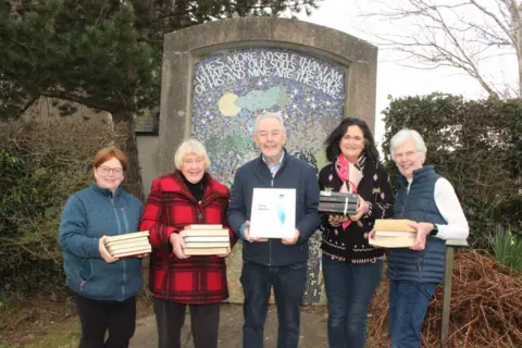 Five people with stacks of books, posing in front of a mosaic of the words "he's more myself than I am. Whatever our souls are made of, his and mine are the same."