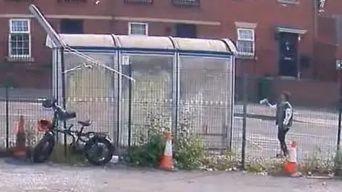 South Yorkshire Police An outdoor area captured by a CCTV camera on 04‑06‑2025 at 15:52:44
A small shelter made of transparent panels stands next to the fence, and a bicycle is parked beside it. Abdullah is visible on the right of the image.