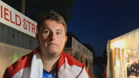 BBC / Zac Sherratt Jamie Power, draped in an England flag, stands outdoors at night near a fan zone, with illuminated stalls and England bunting visible in the background.