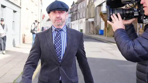 PA Media Terrence McBrien walking past walking past a cameraman in Berwick. He is wearing a tweed cap, jacket, a striped blue shirt and a tie. He has blue eyes and thin greying beard.