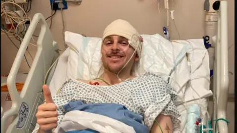 Brain Tumour Research A man in a hospital bed smiling and giving a thumbs up. His head is bandaged and he wears a hospital gown.