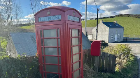 Red telephone box. It has panelled glass walls, a crown emblem at the top with 'telephone' printed below it. A red post box can be seen to the right of it with a small stone building in the distance. Fields can be seen in the background. The sky is blue with some clouds.