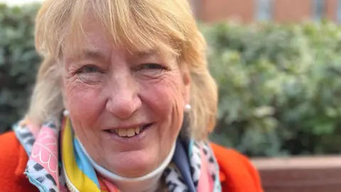 Lady Philippa Dannatt has shoulder-length, blonde hair with red glasses on her head. She is wearing a red jacket with a colourful scarf around her neck and is smiling at the camera.