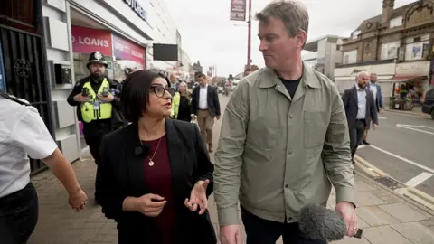 Shabana Mahmood and Ed Thomas walk side by side on a busy high street lined with shops. Ed holds a microphone and appears to be conducting an interview while the other looks toward them while walking. Behind them, several individuals in high-visibility police uniforms and other pedestrians move along the pavement. Shopfronts display signs including “GOLD LOANS,” and cars are parked along the road to the right.