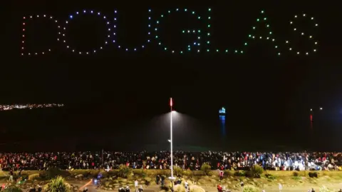 Drones spelling out "Douglas" hover above the bay with their colourful lights a contrast against the black sky behind. Crowds of people fill the promenade walkway below.