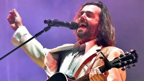 Lord Huron singer Ben Schneider on stage in a white suit with mouth open and eyes closed mid song, with one hand on a guitar neck and the other in the air. At the 2022 Bonnaroo Music & Arts festival in Tennessee.