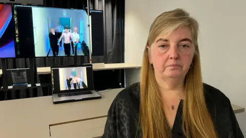 Sharon McFadyen seated indoors in front of a desk, with multiple screens behind showing a video of a shirtless individual being escorted down a corridor by several others; a laptop on the desk mirrors the same footage, and the room includes a white wall, shelving, and dark curtains.