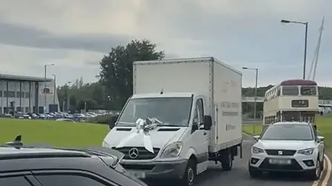 Family photograph A white removals van with a silver bow on the bonnet overtakes a white car and a vintage double-decker bus.