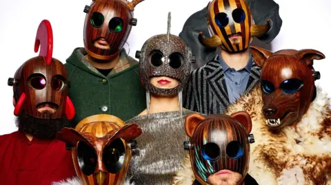 The band FÜLÜ are wearing strange wooden masks, including ones that looks like an owl, a bear and a wolf with big teeth. The band members are standing together and pictured against a white background.
