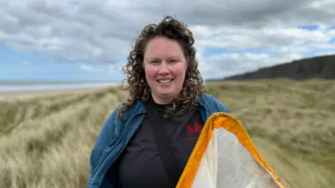 Mel smiling at the camera. She has curly blonde hair and is wearing a grey-black t-shirt with a red Causeway Coast and Glens Heritage Trust logo. She also has a blue jacket and black strap across her chest. Mel is holding a net that has an orange rim. The clouds are overcast and a beach with sand dunes is in the background. 