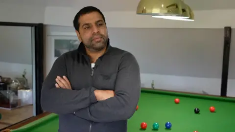 A man in a dark grey zip up jumper is leaning against a pool table with his arms crossed across his chest and is looking towards the right of the camera.