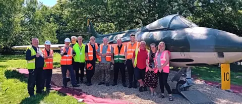 Veterans Central A line of people in hi-vis vests some wearing hard hats celebrate the jet being in its new home.