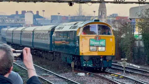 A train pulling into a platform. A man holds a camera and takes a photo.