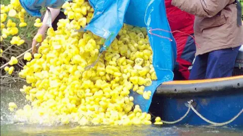 People emptying a large blue sack of hundreds of rubber ducks off the side of a boat and into a river.