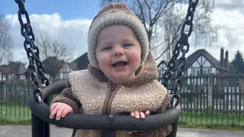 George smiling as he is sitting in a swing. He is wearing a cream bobble hat and beige and brown fleece