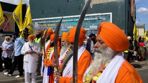 Men in bright orange clothing carrying traditional swords and wearing flower garlands