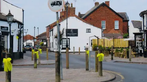 Stand Up For Southport Yellow ribbons tied to wooden bollards in a village street with black and white buildings in the background.