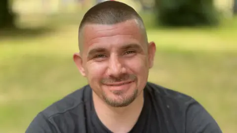 Jack Bennett sonríe a la cámara al aire libre, vistiendo una camiseta de color oscuro.