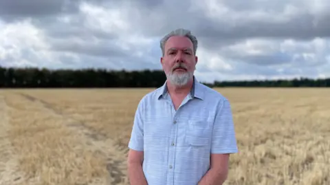 Prof Matthew Goddard - a man wearing a blue short-sleeved shirt - is standing in a harvested field with a woodland in the background under a cloudy sky