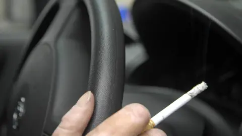 PA Close up of a hand holding a steering wheel and lit cigarette.