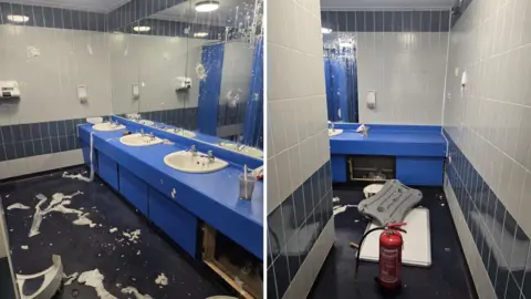 The inside of a public toilet block, there is vandalism of a baby changing table, soap dispensers with wet toilet rolls stuck to walls and mirrors and the floor.