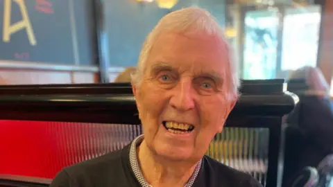 BBC Bill Finlayson looking right at the camera. He has blue eyes and white hair and is smiling. He is wearing a checked shirt under a black jumper. He is sitting in a booth in the Baby Grand and the background behind him is blurred.