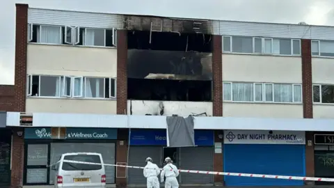 The burnt out flat sits above shops with police tape at ground level and two men in protective suits behind the tape.