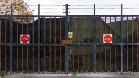 Getty Images Barred gateway to the centre that has metal mesh between the gaps of the bars, barbed wire above it. On each gate there is a red octagonal stop sign. 