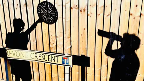 Karen Hurn Shadows of two people are cast onto a wooden fence, with one holding a round bat and the other holding a long object. A street sign reading "Crevone Crescent" stands in the foreground.