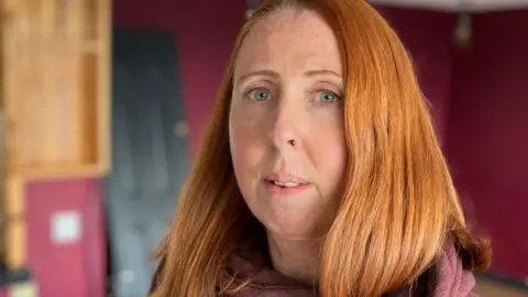 A woman in a red hooded sweatshirt stands inside an empty restaurant. It has no chairs and tables and the bar behind her is empty. She has red, shoulder-length, bobbed hair, blue eyes and freckles, and is looking at the camera.