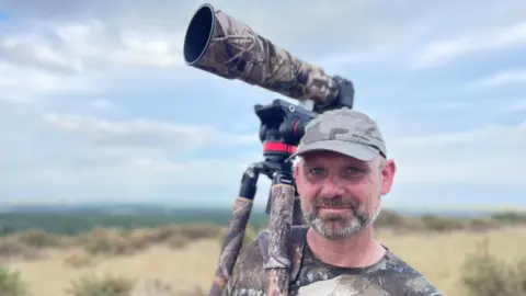 A man with a long lens camera over his shoulder stands at the top of Thoresby Vale Country Park