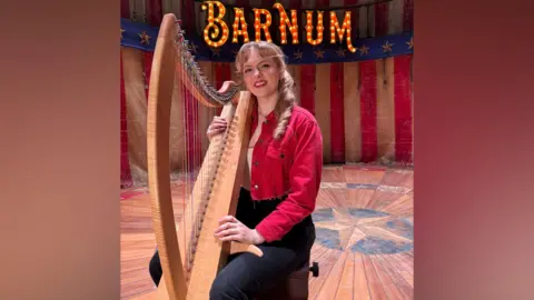 A young woman with dark blonde hair is sitting on a stool on a theatre stage with an American flag type background. She s wearing a red denim cropped jacket, white t-shirt and black jeans and is holding a large light brown harp with colourful strings. On the stage backdrop is the word "Barnum" in large letters lit up by bulbs