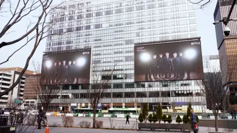 BBC/Jungmin Choi Giant screens mounted on a building at the venue is playing BTS's comeback trailer