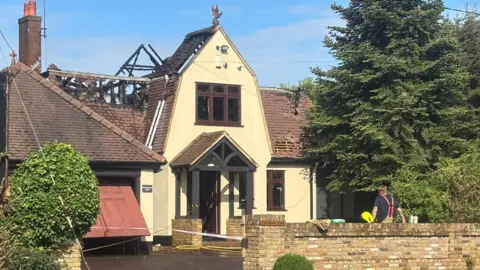 PETER AND ETHAN ROGERS Large home with a beige frontage and brown wooden window frames. You can see part of the roof has been damaged by fire. There's a big fir tree next to the property and a brick wall at the front. The sky is blue and quite clear.