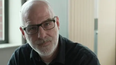 A bald man with a white beard looks into the camera. He is wearing black squared glasses and a black open collared shirt.