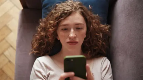A woman lying on a sofa holding her phone up looking bored