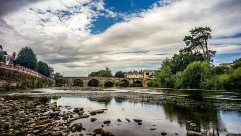 The river bed of the River Wye is seen exposed looking toward Old Bridge after weeks of no rainfall. Buildings and trees can be seen along the right-hand-side of the bank.